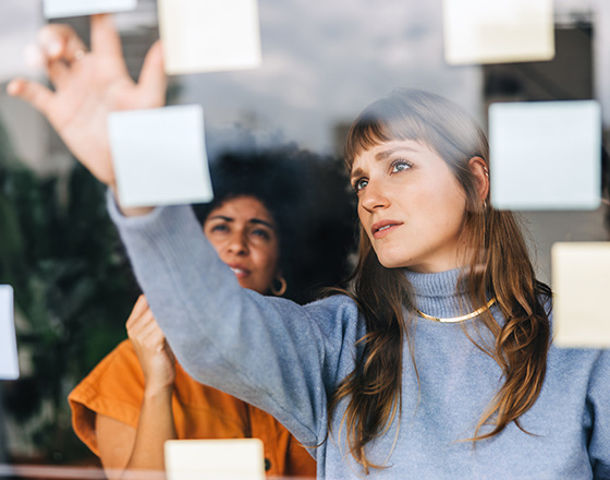 two women brainstorming with sticky notes on glass wall in creative workspace using colorful post its for ideas and collaboration