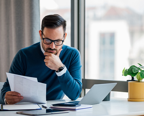 man in glasses sitting at a desk analyzing paperwork with a laptop and notebook in a modern office setting focused on productivity and decision making related to six key aspects of his work
